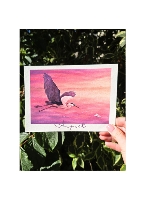 Hand holding a card with a bird illustration and 'August' text against a pink and purple gradient background.