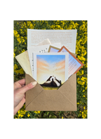 Hand holding a brown envelope with a card featuring a badger and dandelion design against a yellow flower field background.
