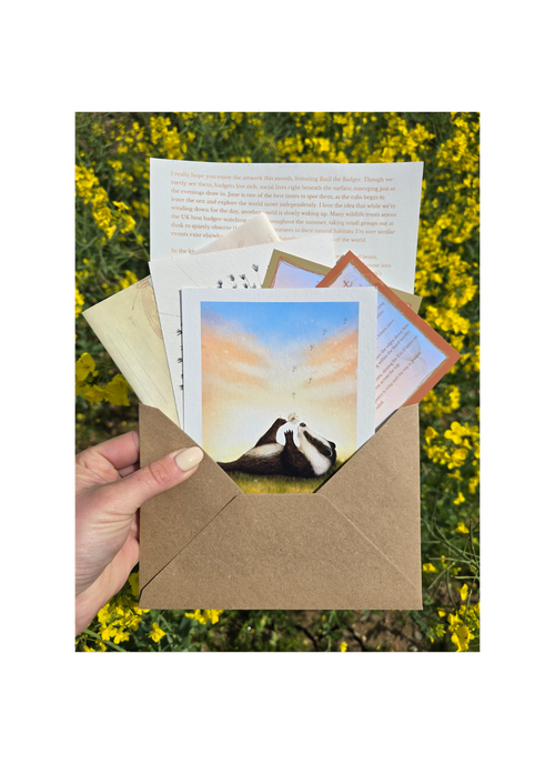 Hand holding a brown envelope with a card featuring a badger and dandelion design against a yellow flower field background.