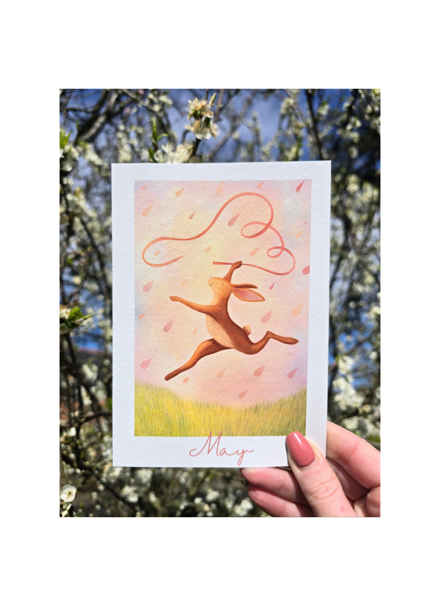 Card with a pink hare illustration and 'May' text held in front of a blurred natural background
