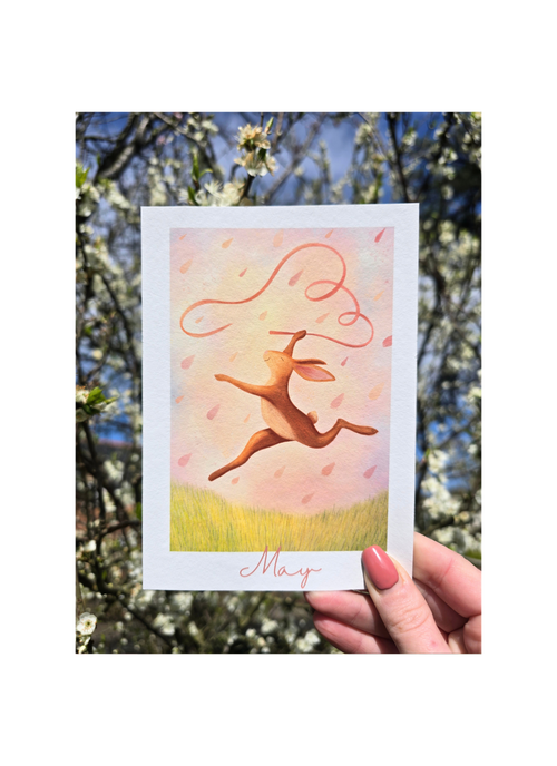 Card with a pink hare illustration and 'May' text held in front of a blurred natural background